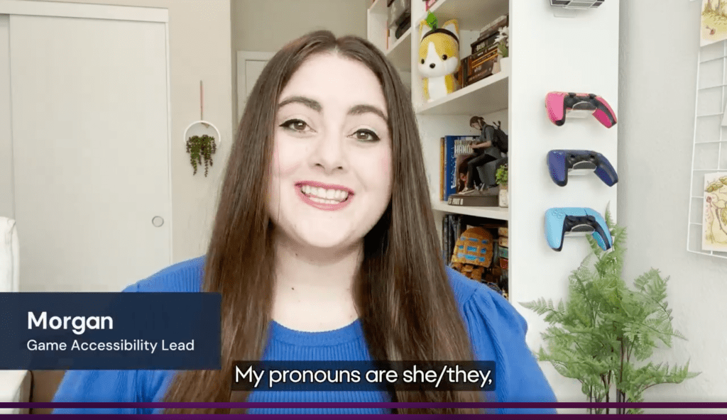 A white woman with long brown hair smiles. Morgan, Game Accessibility Lead. She is stating in the captions, " My pronouns are she/they."